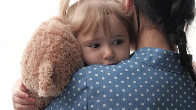 Mutter trägt trauriges Kind auf dem Arm. Kind hält einen Teddybär im Arm.