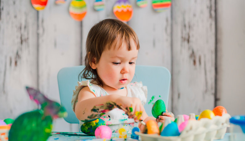 Foto: kleines Mädchen sitzt am Tisch und bemalt Ostereier