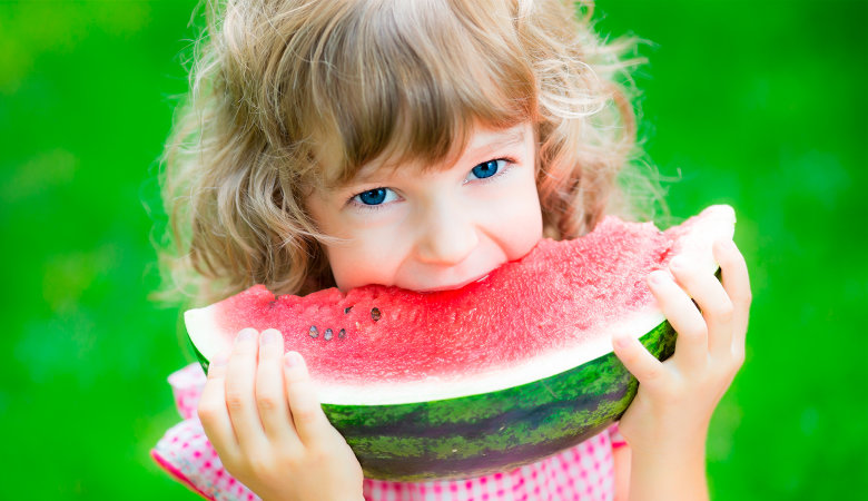 Foto: Kleines Mädchen hält ein großes Stück Wassermelone mit beiden Händen und beißt hinein.