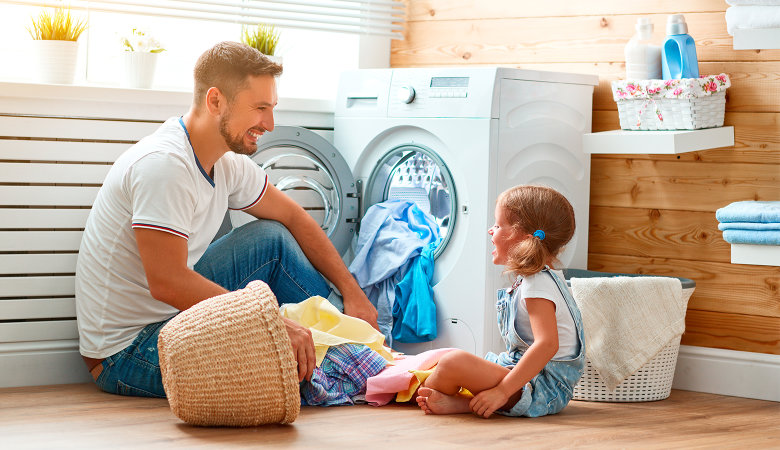 Foto: Fröhlicher Vater und lachende kleine Tochter sitzen vor der geöffneten Waschmaschine am Fußboden