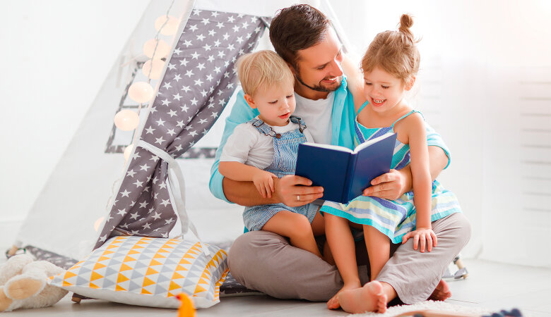 Foto: Vater sitzt mit seinen zwei kleinen Kindern am Fußboden, umarmt die Kinder und liest ein Buch vor