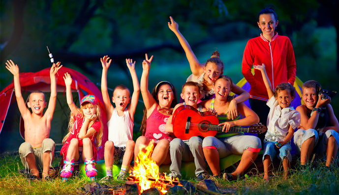 Foto: Neun fröhliche Jugendliche sitzen mit einer Gitarre am Lagerfeuer, eine Erzieherin steht hinter ihnen.