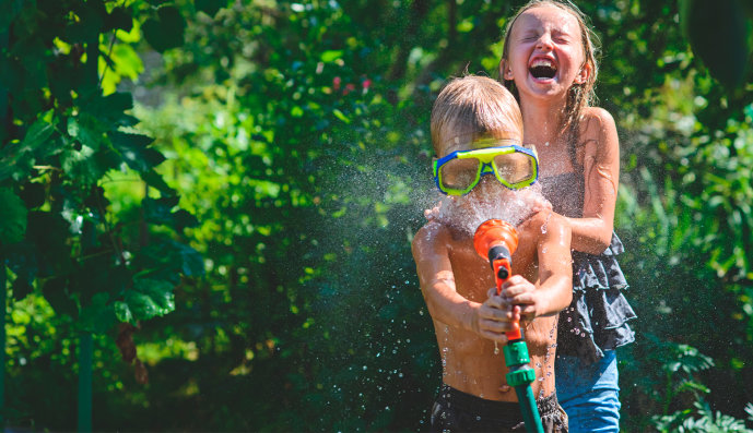 Foto: Zwei lachende Kinder im Garten spritzen sich mit einem Gartenschlauch Wasser ins Gesicht.