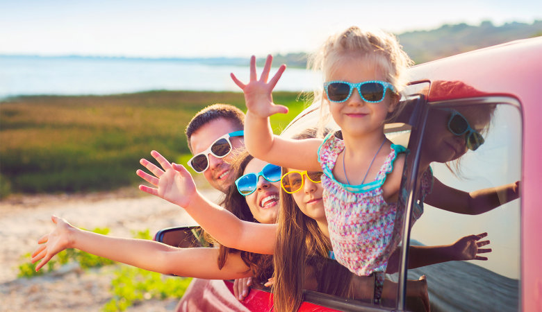 Foto: Familie, Vater, Mutter und zwei Kinder tragen Sonnenbrillen und schauen winkend aus einem Auto raus.