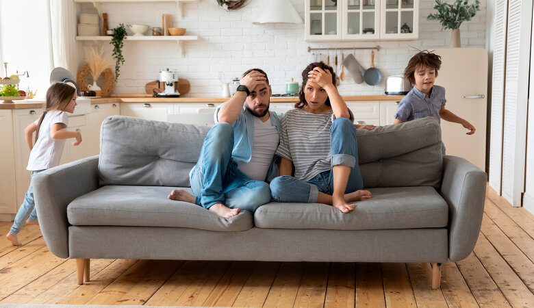 Foto: Gestresstes Paar sitzt auf dem Sofa, Kind tobt um das Sofa herum.