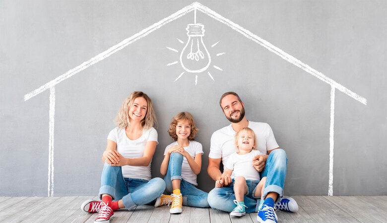 Foto: Familie mit zwei Kindern sitz am Fußboden vor einer Wand auf der die Umrisse eines Hauses gezeichnet sind.