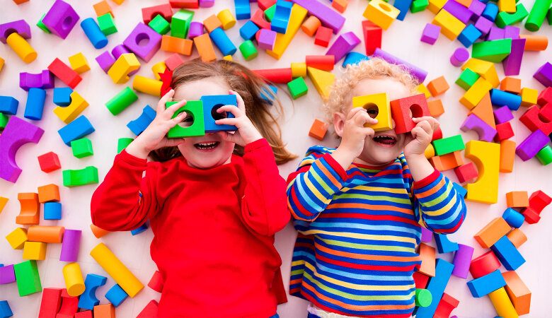 Foto: Zwei kleine Kinder liegen auf dem Rücken auf dem Fußboden und halten sich Plastikspielzeugblöcke 