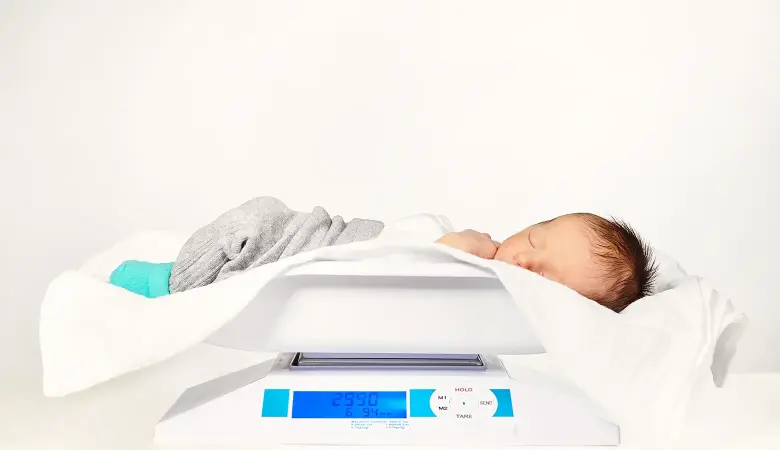Schlafendes Baby liegt auf einer Babywaage. Die Waage zeigt das Gewicht des Babys an.