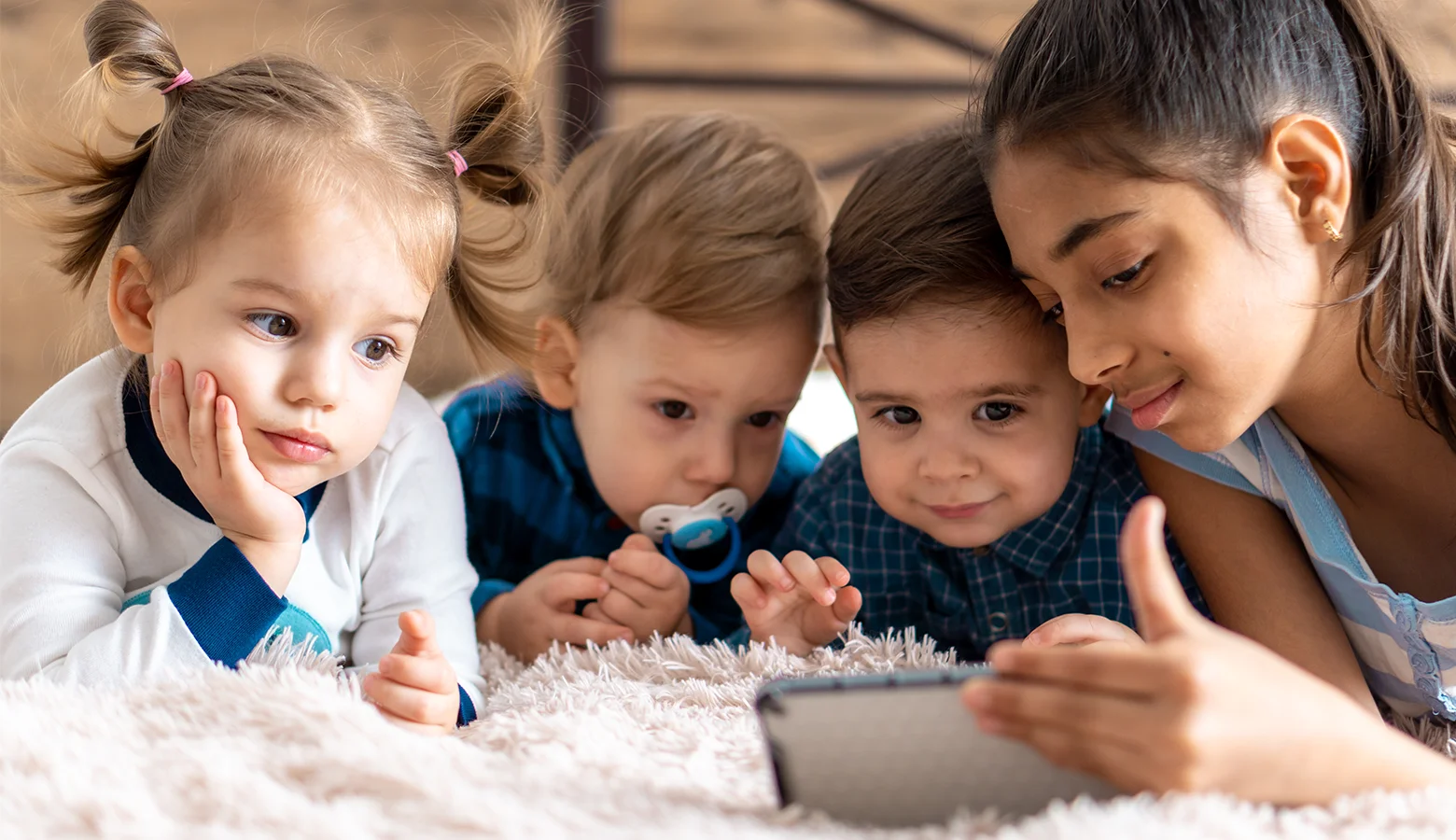 Eine Frau liegt mit drei kleinen Kindern auf dem Teppich und sie schauen in ein Tablet