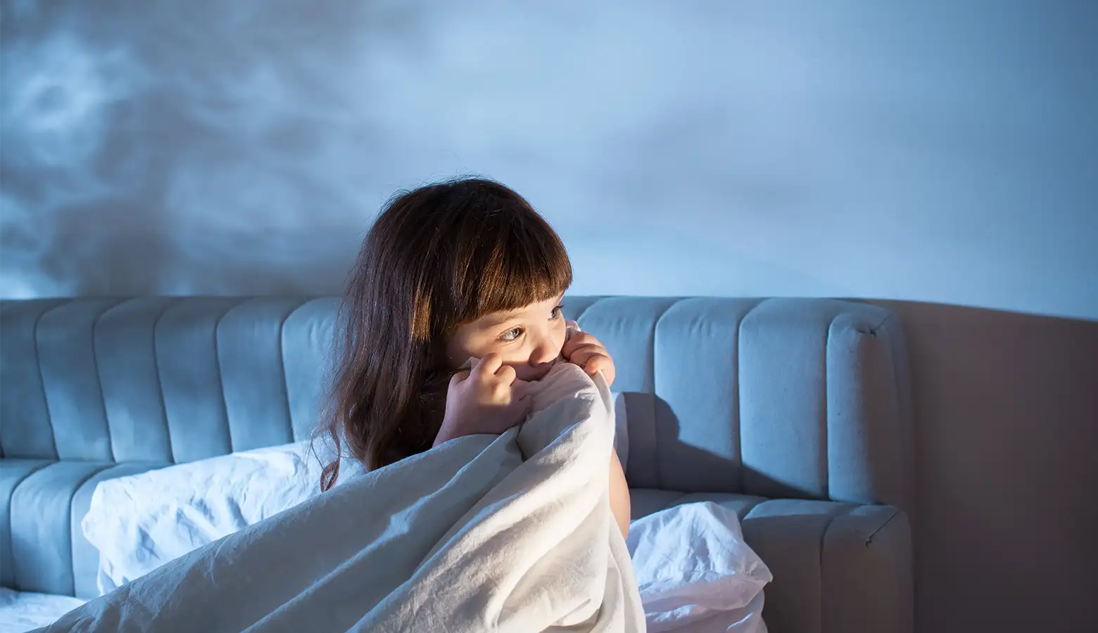 Ein 2 jähriges Mädchen sitzt im Dunkeln auf einem Sofa und versteckt sich unter der Decke, da es Angst hat
