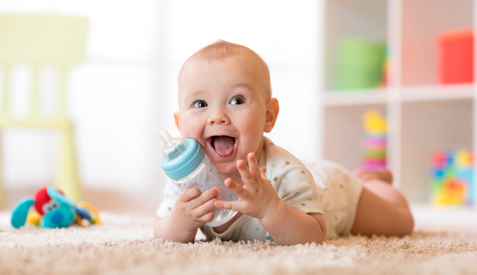 Foto: Ein Baby liegt bäuchlings auf dem Boden. In der Hand hält es eine Milchflasche