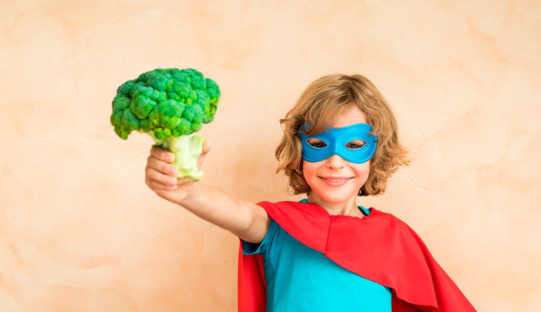 Foto: Junge im  Superhelden Kostüm, hält einen Brokkoli mit ausgestrecktem Arm in Richtung Kamera