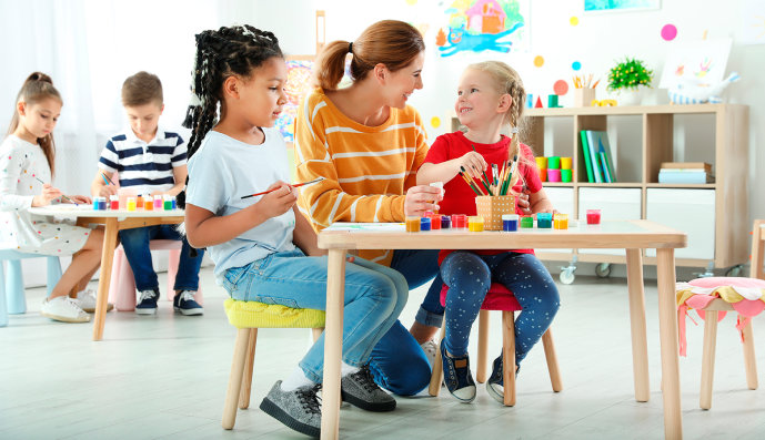 Foto: Kindergärtnerin sitzt mit 2 Kindern an einem Tisch und malt mit diesen