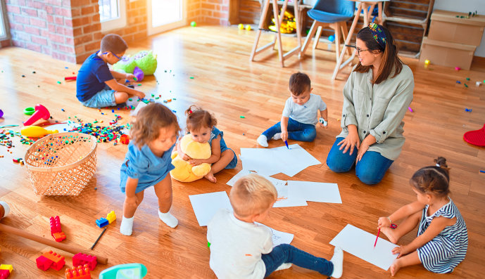 Foto: Kindergärtnerin sitzt mit sechs Kindern auf den Fußboden beim Spielen und Malen.