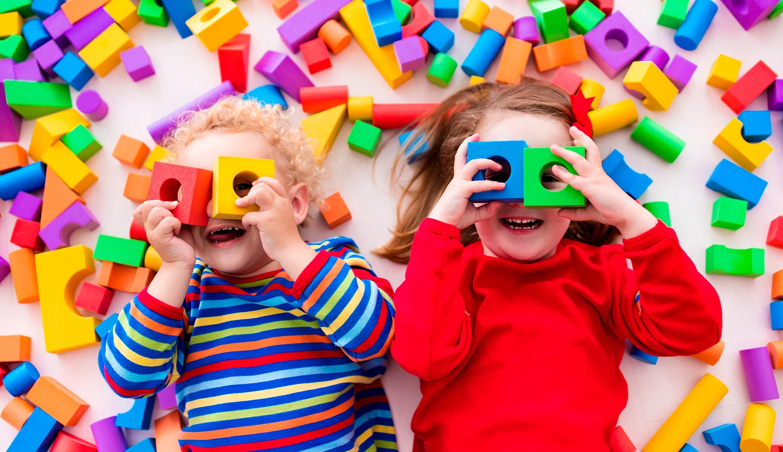 Foto: Zwei kleine Kinder, die auf dem Fußboden liegen und ihre Augen mit Bauklötzen zuhalten.