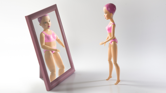 Eine sehr schlanke Barbie-Figur steht vor einem Spiegel. Im Spiegelbild erscheint sie dicker. 