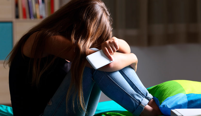 Foto: Trauriges Mädchen mit Handy in der Hand sitzt verzweifelt auf dem Bett.