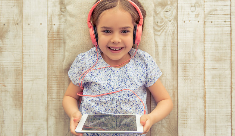 Foto: Kleines Mädchen lacht in die Kamera, hat Kopfhörer auf und ein Tablet in der Hand. 