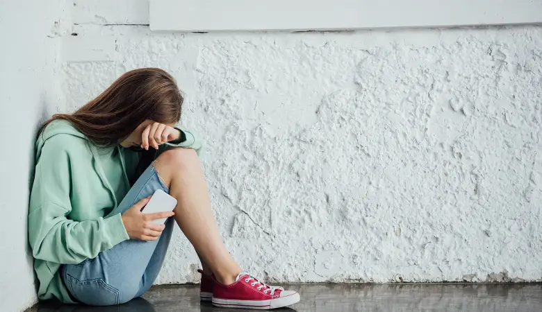 Ein trauriges Mädchen mit Handy in der Hand sitzt an der Wand und hält sich die Augen zu.