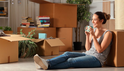 Foto: Junges Mädchen sitzt lachend auf dem Fußboden in ihrer Wohnung mit gepackten Umzugskartons.