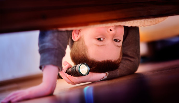 Foto: Kleiner ängstlicher Junge, der mit der Taschenlampe unter das Bett leuchtet.