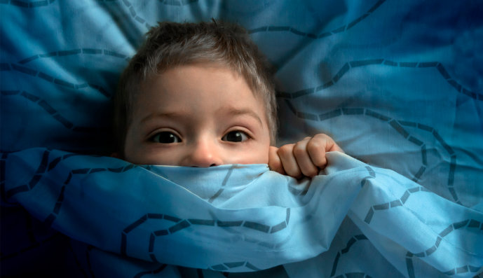 Foto: Junge mit offenen Augen zieht die Bettdecke bis zu Nase hoch und schaut ängstlich.