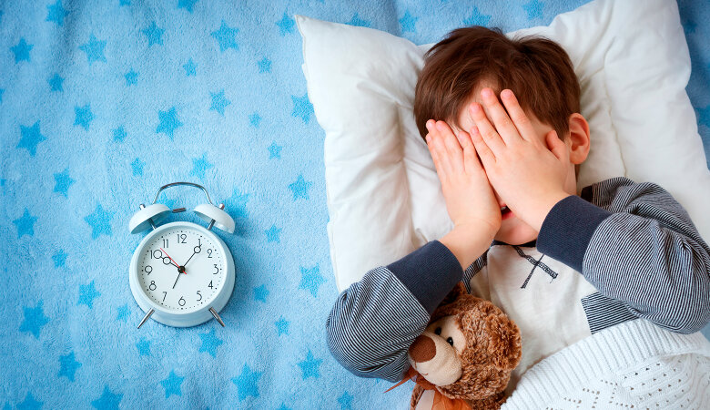 Foto: Sechsjähriger Junge, der mit Teddy und Wecker im Bett liegt und seine Augen zuhält.