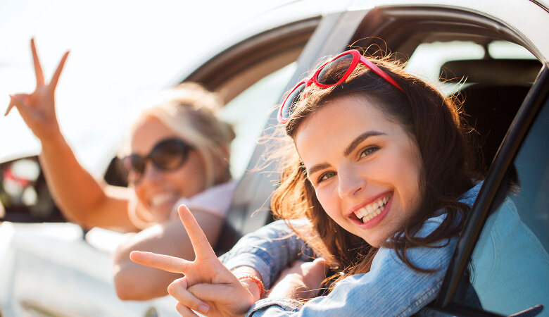 Foto: Zwei lachende Teenagermädchen lehnen sich aus dem Autofenster und winken.