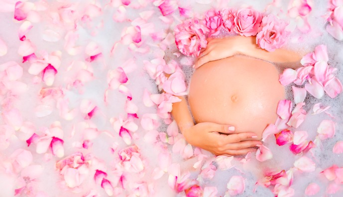 Foto: Schöne schwangere junge Frau, die ihren Bauch in der Badewanne mit Rosenblumen pflegt. 