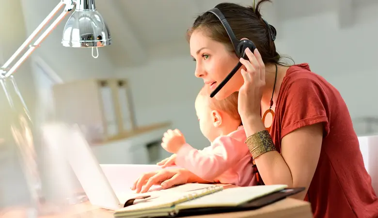 Frau mit Kopfhörer sitzt am Laptop beim Arbeiten und hält Baby im Arm